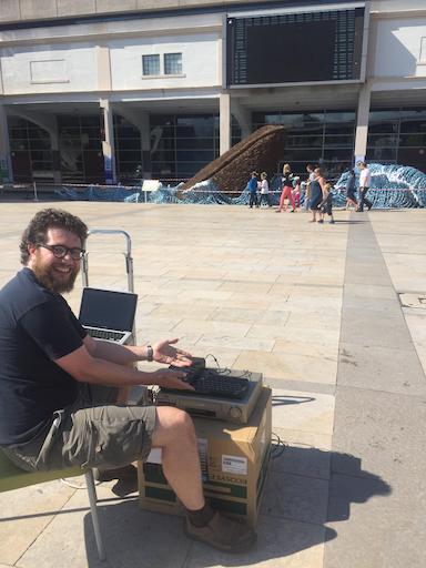 Oliver grinning with a pile of equipment including a ZX Spectrum, a cassette player, a VCR, a UPS and a laptop, while the Spectrum loading screen displays on the Millennium Square Big Screen in the background